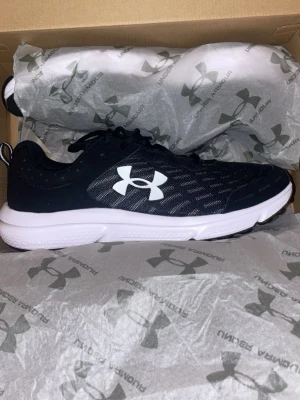 Svarta Under Armour sneakers - Snygga svarta sneakers från Under Armour med vit logga på sidan och vit sula. Ovandelen är i mesh-liknande syntetmaterial för bra andningsförmåga och komfort. Skorna har snörning och en sportig look, perfekta till träning eller vardagsoutfits.