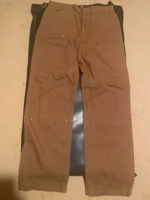 Carhartt WIPDOUBLE KNEE PANT - Tygbyxor - hamilton brown rinsed - Carhartt WIP DOUBLE KNEE PANT - Tygbyxor - hamilton brown rinsed. Byxorna har flera fickor med nitar och hällor för bälte. Tillverkade i slitstark bomull, perfekta för dig som gillar utility-vibe och funktionella detaljer. Helt nya och oanvända. NYPRIS för dessa ligger på 1395 kr 