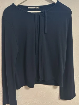 Svart kofta med knyt från 157 - En enkel och stilren svart kofta från 157 med långa ärmar och knyt framtill vid halsen. Perfekt att slänga över en topp eller t-shirt för en chill look. Materialet känns mjukt och lätt, vilket gör den skön att bära hela dagen.