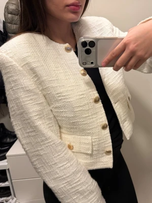 Strukturerad vit kavajjacka från Gina Tricot - Säljer en stilren vit kavajjacka från Gina Tricot. Jackan är croppad med guldiga knappar framtill och på fickorna. Den har en strukturerad, vävd yta och rund halsringning. Perfekt att slänga över en topp för en snygg look. Aldrig använd tyvärr 