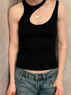 Svart ribbad linnetopp från Zara - Säljer en svart ribbad linnetopp från Zara med asymmetrisk halsringning och ärmlös design. Toppen är figurnära och har en cool cut-out detalj vid halsen som ger en unik look. Perfekt att styla med jeans eller kjol för en trendig outfit.