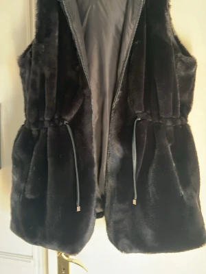 Svart pälsväst från Zhenzi - Mysig svart pälsväst från Zhenzi i storlek M. Västen har dragsko i midjan för snygg passform och stängs med dragkedja framtill. Perfekt att slänga över en hoodie eller tröja för extra värme och stil. Detta är en M i plus size. 