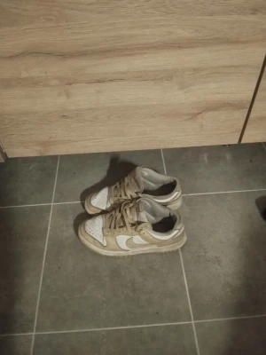 Nike Dunk sneakers beige och vita - Säljer ett par Nike Dunk sneakers i beige och vitt i storlek 40. Skorna har snörning, rund tå och platt sula. perfekta för dig som gillar streetwear och vill ha en clean look.