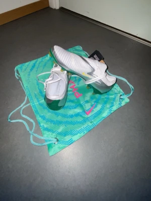 Nike vita fotbollsskor med gröna detaljer - Säljer ett par riktigt snygga vita Nike fotbollsskor med gröna och gula detaljer. Skorna har snörning, en cool silverdetalj på sidan och en skinande grön/gul sula med dobbar. Ovandelen är i syntetmaterial och designen är modern och sportig.