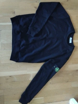 Svart sweatshirt  - Säljer en svart sweatshirt som jag fick av en kompis med klassisk rund hals och ribbade muddar. Tröjan är fästs med knappar. Tillverkad i mjuk bomull för skön känsla och stilren look. Skriv gärna om du har några frågor om tröjan