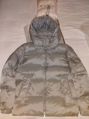 Grå Moncler pufferjacka med huva - Säljer en grå pufferjacka från Moncler med huva och dragkedja framtill. Jackan har två snedställda fickor med dragkedja och Moncler-logga på ärmen. Perfekt för kalla dagar och riktigt snygg till streetwear-stilen.
