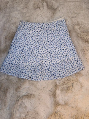 Vit-blå blommig kortkjol - Supersöt kortkjol med vit botten och små blå blommor över hela tyget. Kjolen har en lätt utställd form och känns riktigt somrig och fräsch. Perfekt till en enkel topp och sneakers för en chill look.