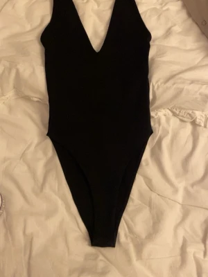 Svart ärmlös bodysuit med v-ringning - Säljer en svart, stilren bodysuit med djup v-ringning och ärmlös design. Perfekt att styla med jeans eller kjol för en clean look. Materialet är stretchigt och följsamt för en bekväm passform. Använt en gång 