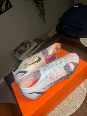 Nike Mercurial vita fotbollsskor - Säljer ett par Nike Mercurial fotbollsskor i vitt med svarta och neonfärgade detaljer. Skorna har snörning, platt sula med dobbar och coola grafiska inslag i rosa, gult och svart. Ovandelen är i syntetmaterial och designen är riktigt sportig och modern.
