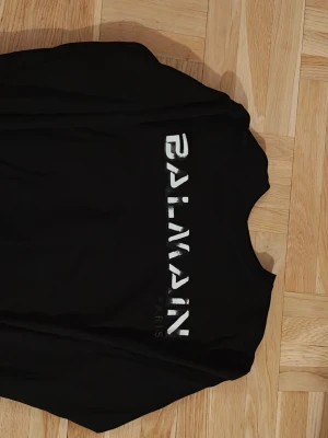 Svart Balmain tröja  - Snygg svart tröja och  har ett stort Balmain-tryck i vitt på framsidan och klassisk rund hals. Perfekt för dig som gillar exklusiva streetwear-vibbar och vill sticka ut med ett ikoniskt märke. Passar för XL tack vare morsan
