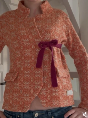 Orange omlottjacka Odd Molly - Unik orange omlottjacka från Odd Molly med vitt geometriskt mönster och lila rosett i midjan. Jackan har hög krage, två fickor fram och ett snyggt broderat märke på ärmen. Tillverkad i mjuk ullblandning och har lång ärm.🤩💕                                             Hör gärna av er om det behövs fler bilder!😊