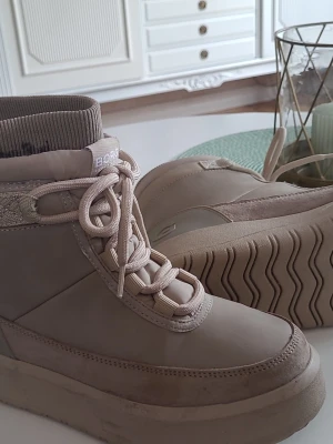 Beige boots från Björn Borg - Snygga beige boots från Björn Borg med högt skaft och tjock sula. Skorna har snörning framtill, ribbad mudd upptill och är gjorda i en mix av mocka och syntet. Perfekta för höst och vinter när du vill ha både stil och komfort. Storlek 38, en aning små för att vara 38.