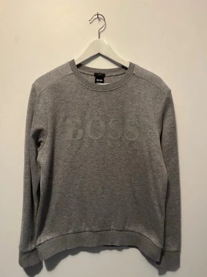 Grå sweatshirt från Hugo Boss - Säljer en stilren grå sweatshirt från Hugo Boss med stor BOSS-logga framtill. Tröjan har rund halsringning, långa ärmar och mjukt material i bomullsblandning. Perfekt för en avslappnad look och enkel att matcha med jeans eller joggers.