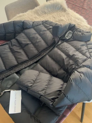 Svart pufferjacka från Calvin Klein - Säljer en svart pufferjacka från Calvin Klein. Jackan har dragkedja, hög krage och är lätt vadderad för extra värme. Perfekt för kyliga dagar och riktigt snygg till streetwear.
