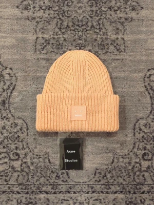 Mössa från acne studios - Säljer en stickad mössa från Acne Studios med det ikoniska ansiktsloggan framtill. Mössan är ribbstickad och gjord i 100% ull, vilket gör den både varm och snygg. Perfekt accessoar för att lyfta din outfit under kyliga dagar.