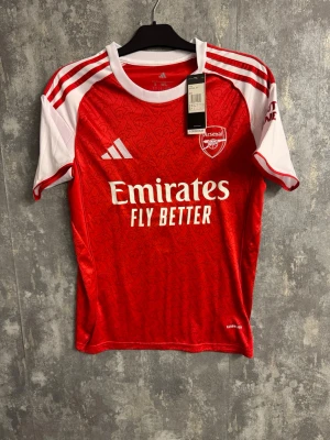 Arsenal matchtröja Saka 7 Adidas - Röd och vit Arsenal matchtröja från Adidas med Saka 7 på ryggen. Tröjan har vita detaljer på ärmarna, klassiska Adidas-ränder och klubbmärke på bröstet. Tillverkad i lätt polyester med diskret mönster i tyget och Aeroready-teknologi.