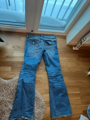 Blå bootcut jeans med slitningar - Säljer ett par ljusblå bootcut jeans med slitna detaljer och hål på framsidan och snygga fickor med knapp ibak. 