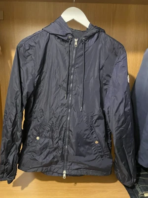 Mörkblå vindjacka från Moncler - Snygg mörkblå vindjacka från Moncler med huva och dragsko. Jackan har dragkedja framtill, två fickor med tryckknappar och Moncler-logga på ärmen. Tillverkad i lätt polyester som passar perfekt för blåsiga dagar.