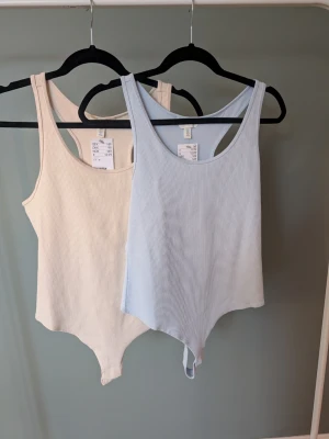 Ribbad ljusblå och beige linnetopp - Två ribbade linnetoppar från H&M i storlek M, en i ljusblå och en i beige. Båda har rundad hals och brottarrygg, tillverkade i mjuk bomullsmix. Perfekt för en chill och enkel stil, passar till jeans eller shorts.