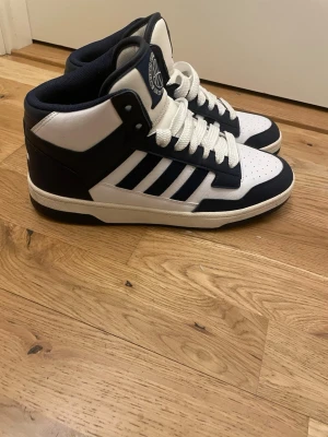 Adidas sneakers high-top marinblå/vita - Säljer ett par snygga high-top sneakers från Adidas i marinblått och vitt. Skorna har klassiska tre ränder på sidan, perforerad tå och tjock vit sula. Tillverkade i syntetmaterial med skön vadderad insida och snörning framtill. Perfekta för dig som gillar retro basketstil.