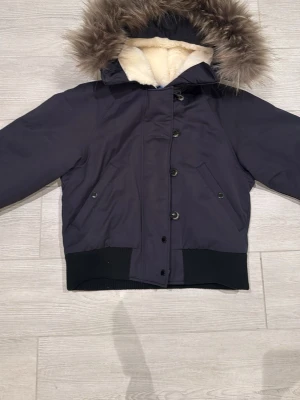 Svart vinterjacka Peak Performance - Säljer en svart vinterjacka från Peak Performance med mysig fuskpäls på huvan och blått quiltat foder. Jackan har knappar och dragkedja framtill, två fickor med lock och ribbade muddar. Perfekt för kalla dagar och riktigt snygg streetstil.