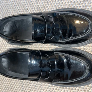Svarta chunky loafers i lack - Säljer ett par svarta loafers med chunky sula och glansig lackyta. Skorna har klassisk pennyloafers-design med rund tå och platt sula. Perfekta för dig som vill ha en edgy men stilren look. Storlek 37.