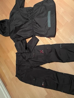 Hundförare- Svart skalställ High Mountain - Säljer ett svart skalställ från Engelson med jacka (36) och byxor(38). Jackan har huva, dragsko i midjan och flera fickor med knappar, ficka i ryggen för dummy eller andra hundsaker.. Byxorna har praktiska fickor och detaljer i rött. Perfekt för hundträning i ur och skur. Pris för både byxor och jacka 500. 