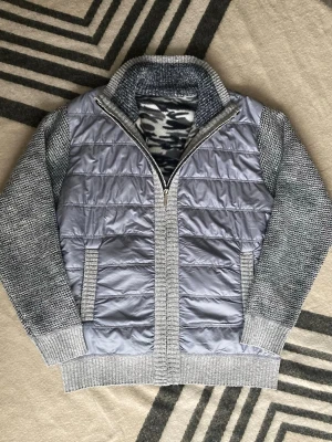 Grå quiltad cardigan från Fashton BB - Snygg grå cardigan jacka med quiltad framsida i blankt material och stickade ärmar samt rygg. Jackan har ribbade muddar, hög krage och dragkedja framtill. Insidan har ett coolt camouflage-mönster vid kragen. Perfekt för dig som gillar lager på lager.