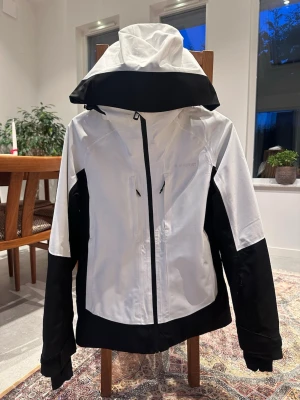 Snygg skidjacka från Everest - Säljer en svartvit skidjacka från Everest med huva och dragkedja framtill. Jackan har flera praktiska fickor med dragkedjor och justerbara muddar vid ärmsluten. Perfekt om man är ute i backen. Den har även en ficka för liftkort.