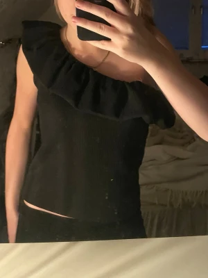 Svart oneshoulder topp med volang - Säljer en svart oneshoulder topp med bred volang längs halsringningen. 