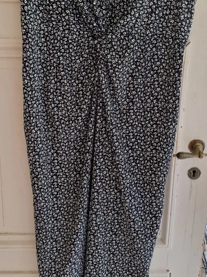 Svart blommig byxdress - Säljer en svart byxdress med små vita blommor och knyt framtill. Västen har en figurnära passform och är perfekt för att lyfta en enkel outfit. Materialet känns mjukt och följsamt, troligen polyester eller bomull. Djur finns i hemmet.