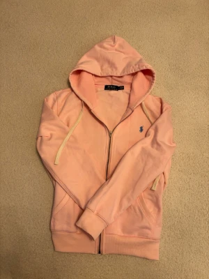 Polo Ralph Lauren rosa hoodie - En mjukt rosafärgad hoodie som utstrålar en avslappnad men stilren elegans. Den har en klassisk dragkedja framtill, en bekväm huva med ljusa snören och ribbade muddar som ger en fin passform. Det diskreta, broderade Polo Ralph Lauren-emblemet på bröstet adderar en tidlös och exklusiv känsla. Tyget ser mjukt och inbjudande ut – perfekt för både lugna hemmadagar och lediga utflykter, där komfort möter genomtänkt design.