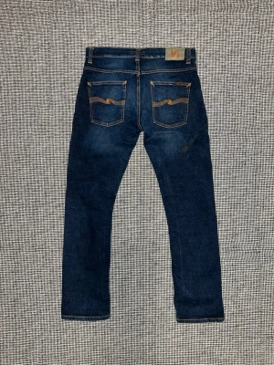 Nudie Jeans Grim Tim blå jeans 32/30 - Säljer ett par Nudie Jeans Grim Tim i modellen Authentic Deep. Mörkblå jeans med snygga slitningar och klassiska orange sömmar. Tillverkade av 99% ekologisk bomull och 1% elastan för lite stretch. Rak passform och ikoniska bakfickor med vågig söm.