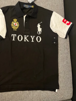 Polo Ralph Lauren T-shirt (ny)  - Säljer en svart pikétröja från Polo Ralph Lauren med vita ärmar och stora broderade detaljer framtill, inklusive Tokyo-text, logga och emblem. Klassisk krage och knappar vid halsen. Röd siffra på ena ärmen ger extra sportig känsla. Passar även storlek M