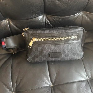 Gucci Midjeväska - Säljer en äkta Gucci midjeväska (belt bag) i svart med justerbart bälte och klassisk röd/blå Web-rand samt silverfärgad dragkedja.  Autentiserad av Tradera (officiell äkthetskontroll) Kvitto finns Serienummer finns  Väskan är i bra skick. Det finns ett mycket diskret slitage på en liten del av bandet, inget som påverkar användning eller helhetsintryck (se bild).  Fraktas spårbart och försäkrat. Skickas snabbt efter betalning.  Vid seriöst intresse – skicka gärna meddelande.