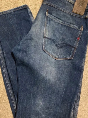 Replay Anbass blå jeans, storlek 33 - Säljer ett par Replay Anbass jeans i klassisk blå tvätt. Jeansen har snygga slitningar, raka ben och detaljer som orange söm vid hällan och Replay-logga på bakfickan. Tillverkade i slitstarkt denim med fem fickor och knappgylf.