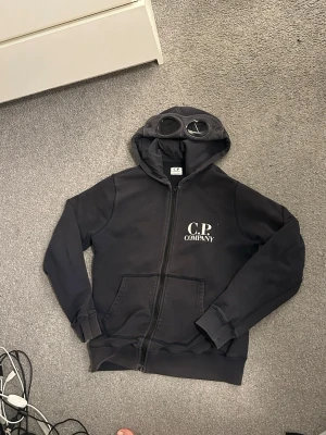 Cp company tröja med glasögon  - VID SNABB AFFÄR = BILLIGARE PRIS! Detta är en cp company tröja. Den är äkta, har bevis i fotot. Den är i storlek 12 står det. Den har glasögon i luvan. Den är lite solblekt men går att fixa. Den är ganska mörkblå går med åt det svarta hålet. Den är perfekt för dig som gillar tröja med dragkedja. Den har ett pytte pytte litet hål i armhålan(märks inte) SÄLJER BILLIGT FÖR JAG VILL HA BORT DEN (den ligger bara och inte används för den är för liten för mig) 