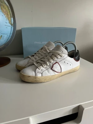 Vita Philippe Model sneakers med röd detalj - Säljer ett par vita Philippe Model sneakers i skinn med klassisk låg profil och rund tå. Skorna har snörning, svart hälparti och en röd kantad sköld på sidan. Sulan är beige och ger en vintagekänsla. Perfekta för dig som gillar stilrena men ändå unika sneakers.