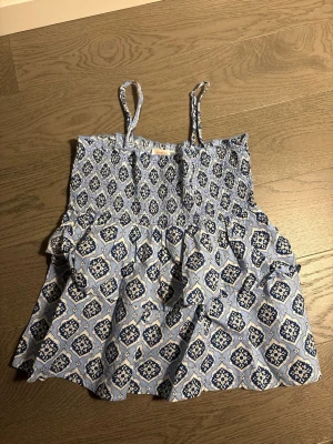 Blå mönstrad topp/ blus från Gina Tricot - Supersöt blå och vit topp från Gina Tricot med smala axelband och smock upptill. Toppen har ett detaljerat mönster i blått och vitt och är gjord i ett lätt tyg, perfekt för varma dagar. 💙💙aldrig använt, storlek 146/152-xxxs
