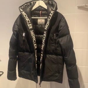 Svart dunjacka från Moncler Short Down - Snygg svart dunjacka från Moncler med logotyp längs dragkedjan. Jackan har en quiltad design och är perfekt för kyligare väder. Den har en huva och praktiska fickor med dragkedja. Perfekt för att hålla stilen och värmen under vintern.