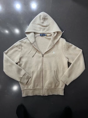Beige zip Hoodie från Polo Ralph Lauren - Snygg beige hoodie från Polo Ralph Lauren med broderad logga på bröstet. Tröjan har huva med snörning, två fickor fram och hel dragkedja med Polo-detalj. Tillverkad i mjuk bomullsmix, perfekt för chill dagar. Klassisk och stilren design. Zip hoodien är äkta och införskaffades för cirka ngn månad sedan och är fortfarande i strålande skick.
