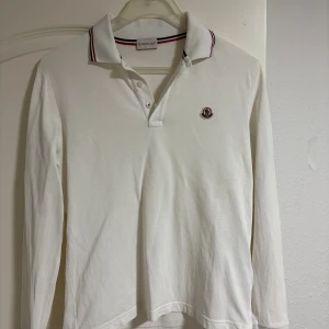 Vit långärmad pikétröja Moncler - Vit långärmad pikétröja från Moncler med klassisk krage och knäppning framtill. Tröjan har Moncler-logga broderad på bröstet och snygga ränder i rött och svart på kragen. Tillverkad i mjuk bomull som känns skön mot huden. Storlek M men väldigt liten i storlek, passar S eller xs 