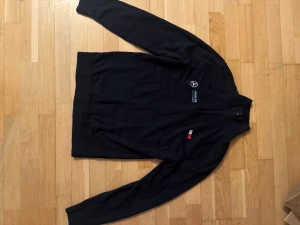 Svart half zip tröja Tommy Hilfiger AMG - Svart långärmad tröja från Tommy Hilfiger x Mercedes AMG Petronas med half zip och broderade loggor på bröstet. Tröjan har en stilren design med ribbade muddar och klassisk passform. Perfekt för dig som gillar motorsport och snygga samarbeten.
