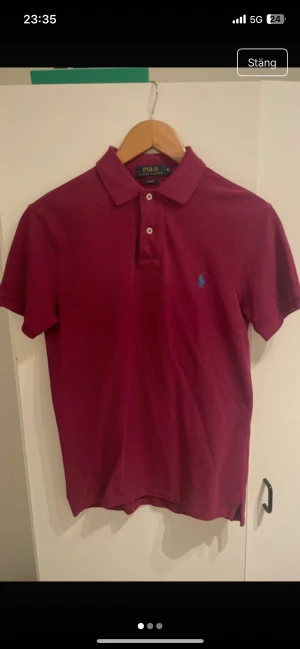 Vinröd pikétröja från Ralph Lauren - Snygg vinröd pikétröja från Ralph Lauren med klassisk krage och två knappar framtill. Tröjan har korta ärmar och den ikoniska blå logon broderad på bröstet. Perfekt för dig som gillar stilrena och tidlösa plagg.