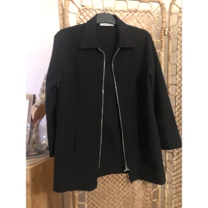 Svart kavaj med dragkedja från Lindex - Stilren svart kavaj från Collection by Lindex i storlek 38/40. Kavajen har en klassisk krage och en snygg silverfärgad dragkedja framtill som ger en modern touch. Passar perfekt till dig som gillar minimalistisk och clean stil. Perfekt till nyår!