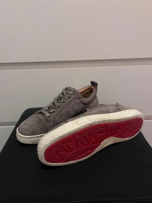 Gråa Louboutin skor - Snygga grå sneakers i mocka från Louboutin med den ikoniska röda sulan. Skorna har rund tå, platt vit sula och finns med både gråa och vita skosnören.  Perfekt om du vill ha det kul i vinter ❄️ 