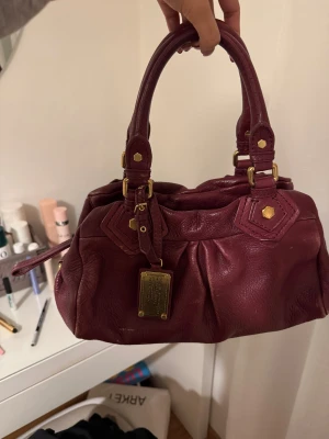 Marc Jacobs väska - Snygg vinröd handväska från Marc Jacobs i äkta skinn med guldiga metalldetaljer och klassisk logga framtill❤️