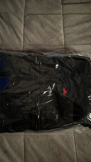 Svart hoodie från Polo Ralph Lauren - Svart hoodie från Polo Ralph Lauren med dragkedja framtill och klassisk röd broderad logga på bröstet. Hoodien har huva och är tillverkad i mjukt bomullsmaterial, perfekt för en chill och stilren look.