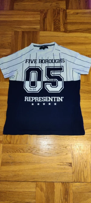 Randig t-shirt Outfitters Nation S - T-shirt i baseballstil från Outfitters Nation med svart och vit färgblock, tunna svarta ränder upptill och tryck med text och siffror framtill. Kortärmad och i mjuk bomull, perfekt för en sportig streetlook.