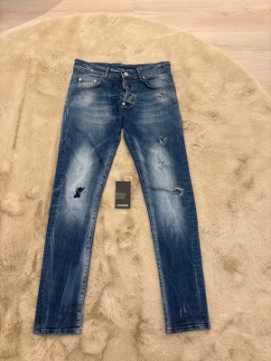  jeans från Dsquared2 - Snygga blå jeans från Dsquared2 med slitningar, färgstänk och skinny passform. Klassisk femficksmodell med låg midja och tydlig Dsquared2-logga på bakfickan. Jeansen har dragkedja och knapp framtill, samt coola detaljer som ger en edgy vibe. Kom dm för eventuella frågor 🧐pris kan diskuteras 💯. Ops vissa av slitningarna har blivit till hål